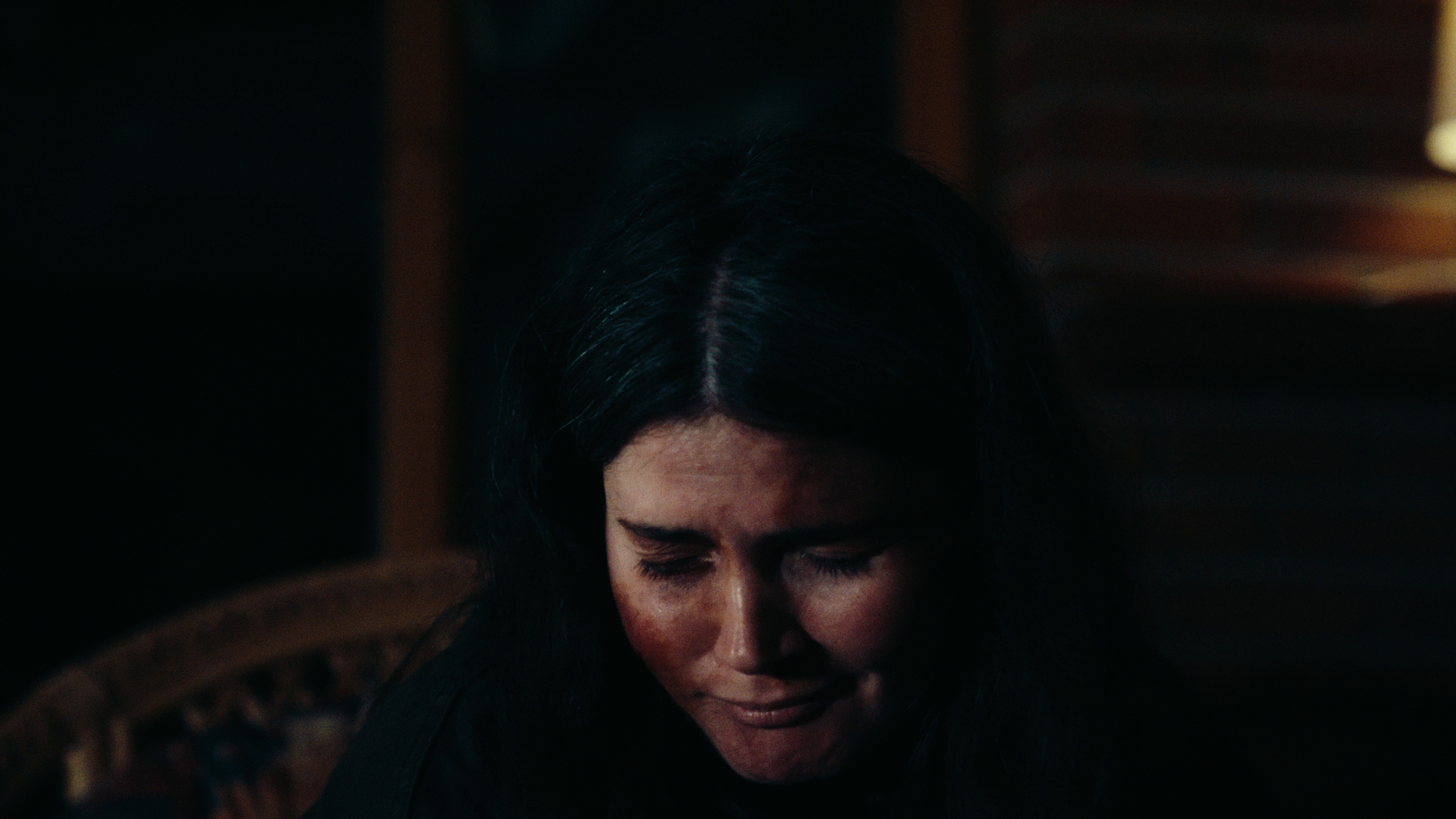 Close-up of a woman with dark hair and a contemplative expression, sitting in a chair in a dimly lit room.