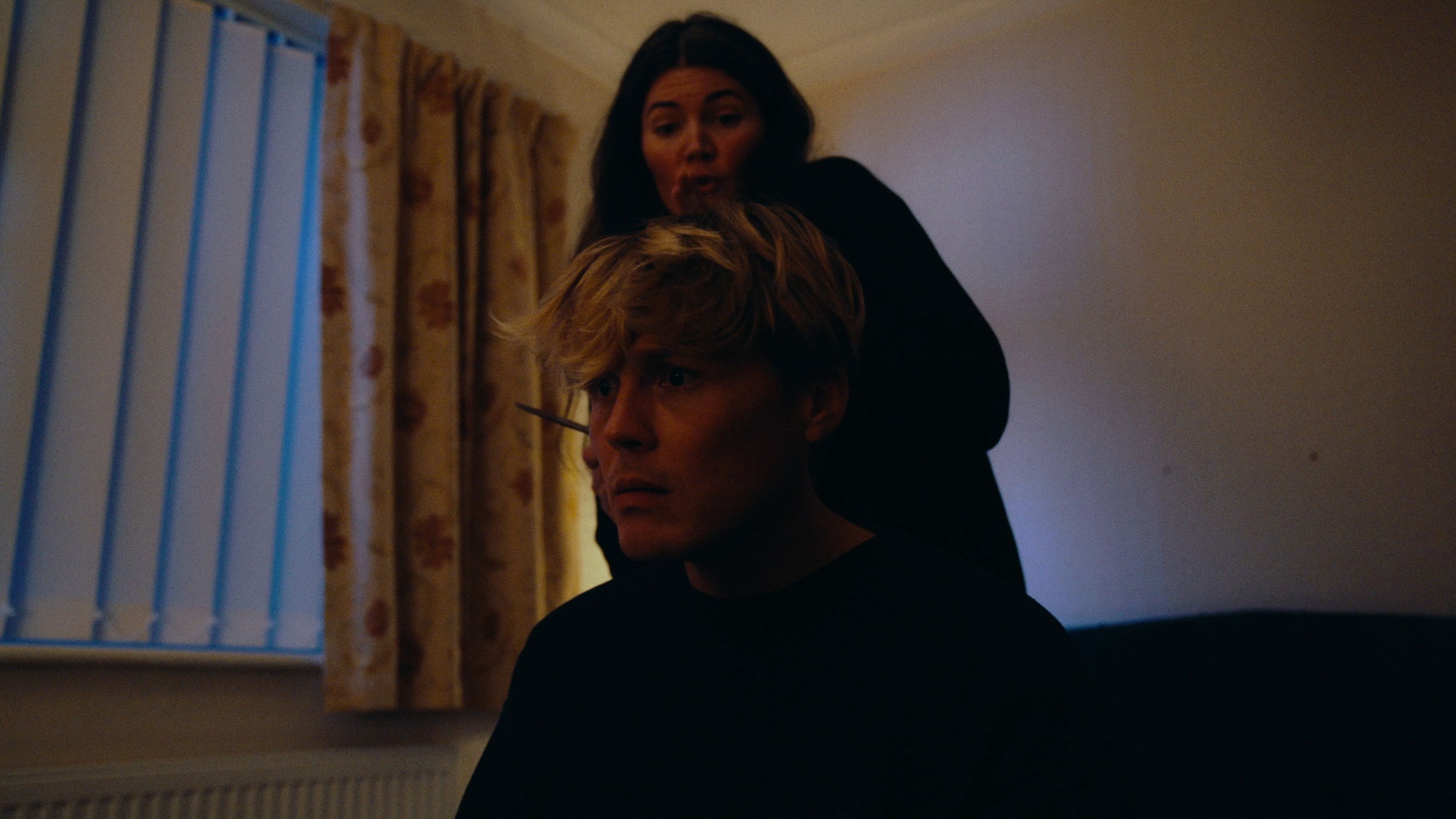 A woman with long dark hair looking down at a young man with blonde hair, who has a pencil in his ear, in a dimly lit room with striped curtains.