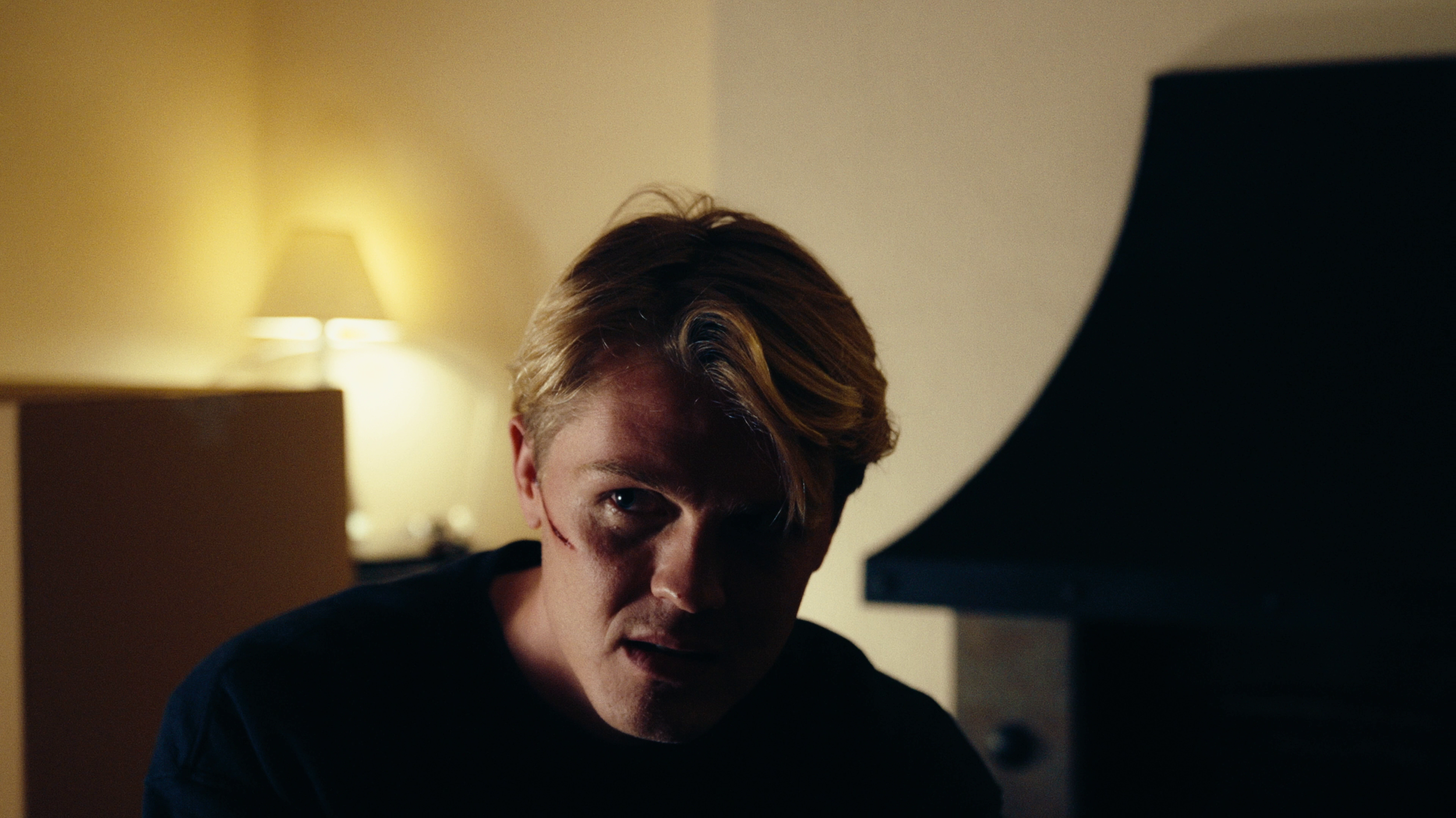 A young man with red hair and a scratch on his face is sitting in front of a computer in a dimly lit room, illuminated by a lamp on a desk.
