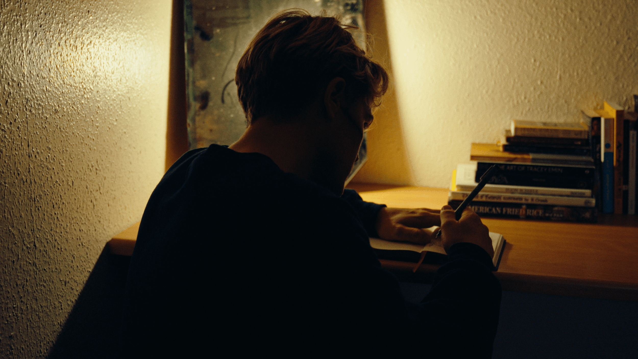 A person with short hair is sitting at a desk, looking at a cell phone while writing in a notebook. There is a small bookshelf with various books on the right side of the desk, and a mirror is visible in the background behind the person.