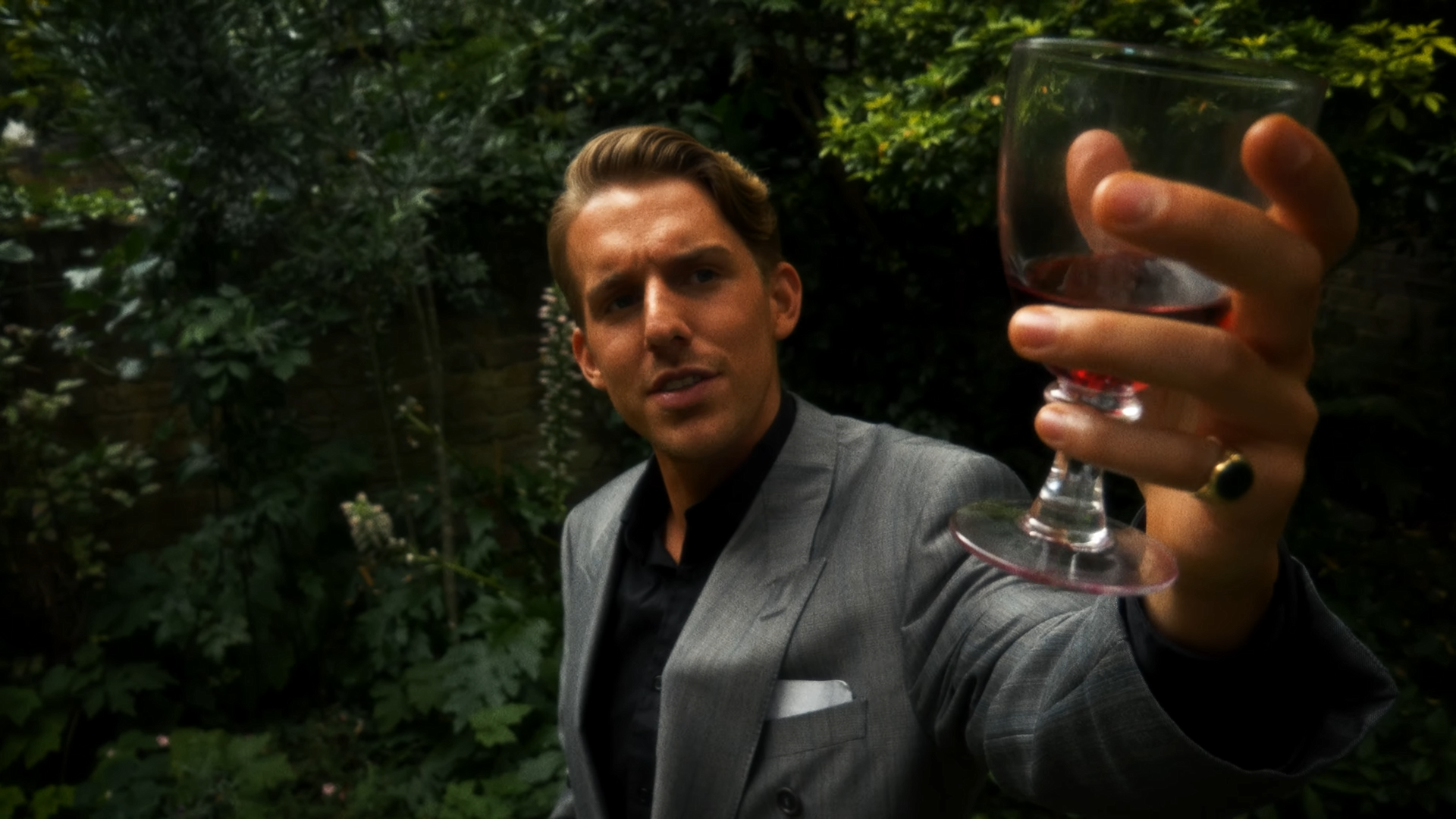 Man in a gray suit and black shirt holding a glass of red wine, raising it in a toast outdoors with green foliage in the background.