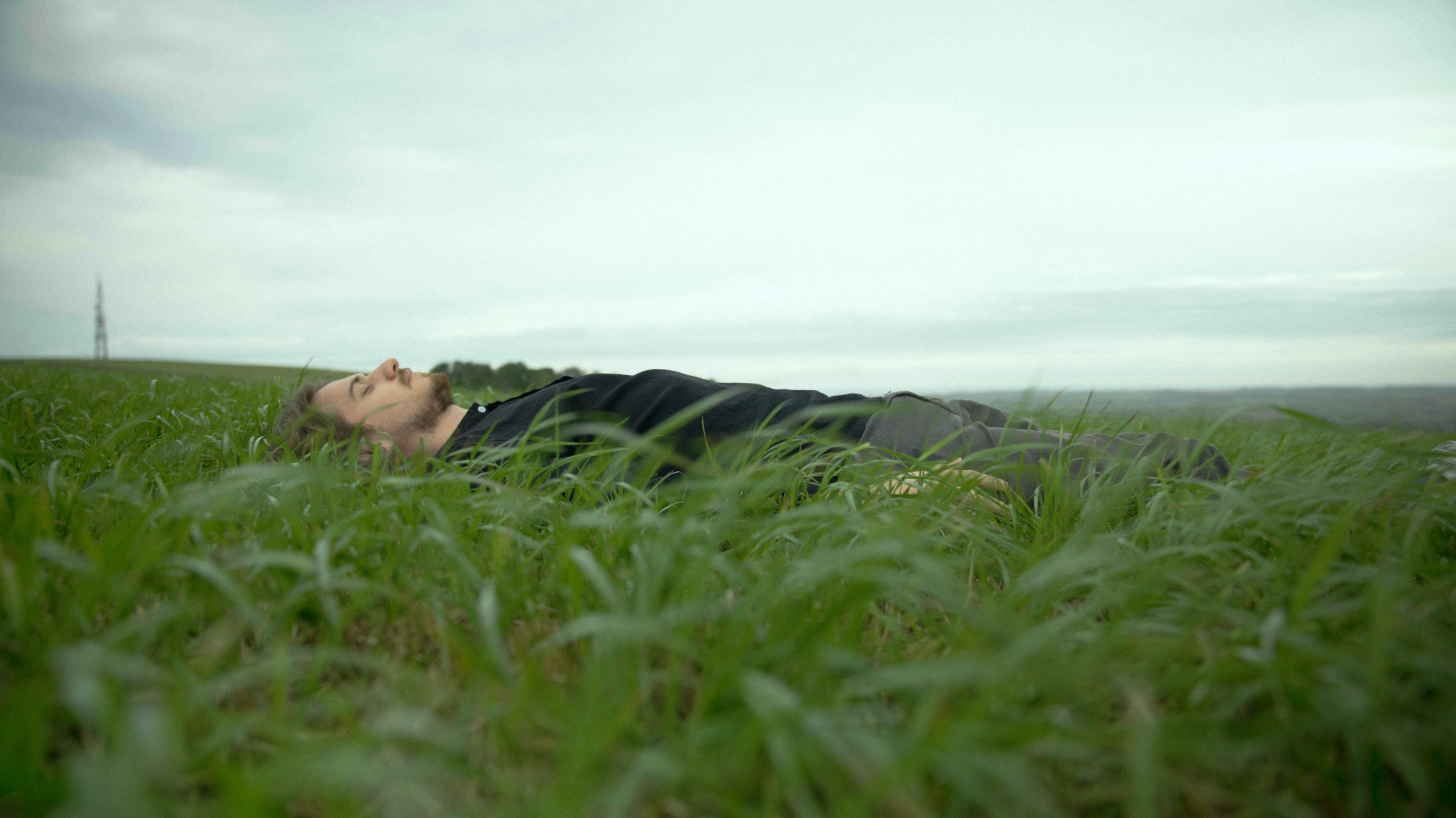 A man lying down in a grassy field, looking relaxed, with an overcast sky.