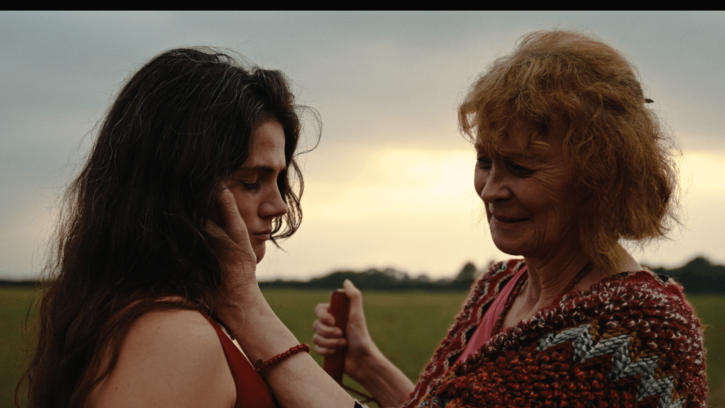 A young woman with long dark hair and a red dress is close to an older woman with curly red hair and a colorful knitted sweater, both smiling and touching each other's faces outdoors at sunset.