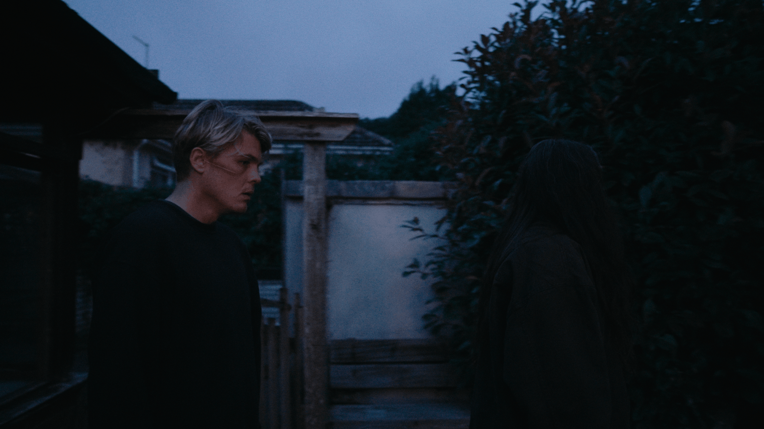 A young man with blond hair standing outdoors near a hedge, with another person with long dark hair facing away, during dusk or early evening.