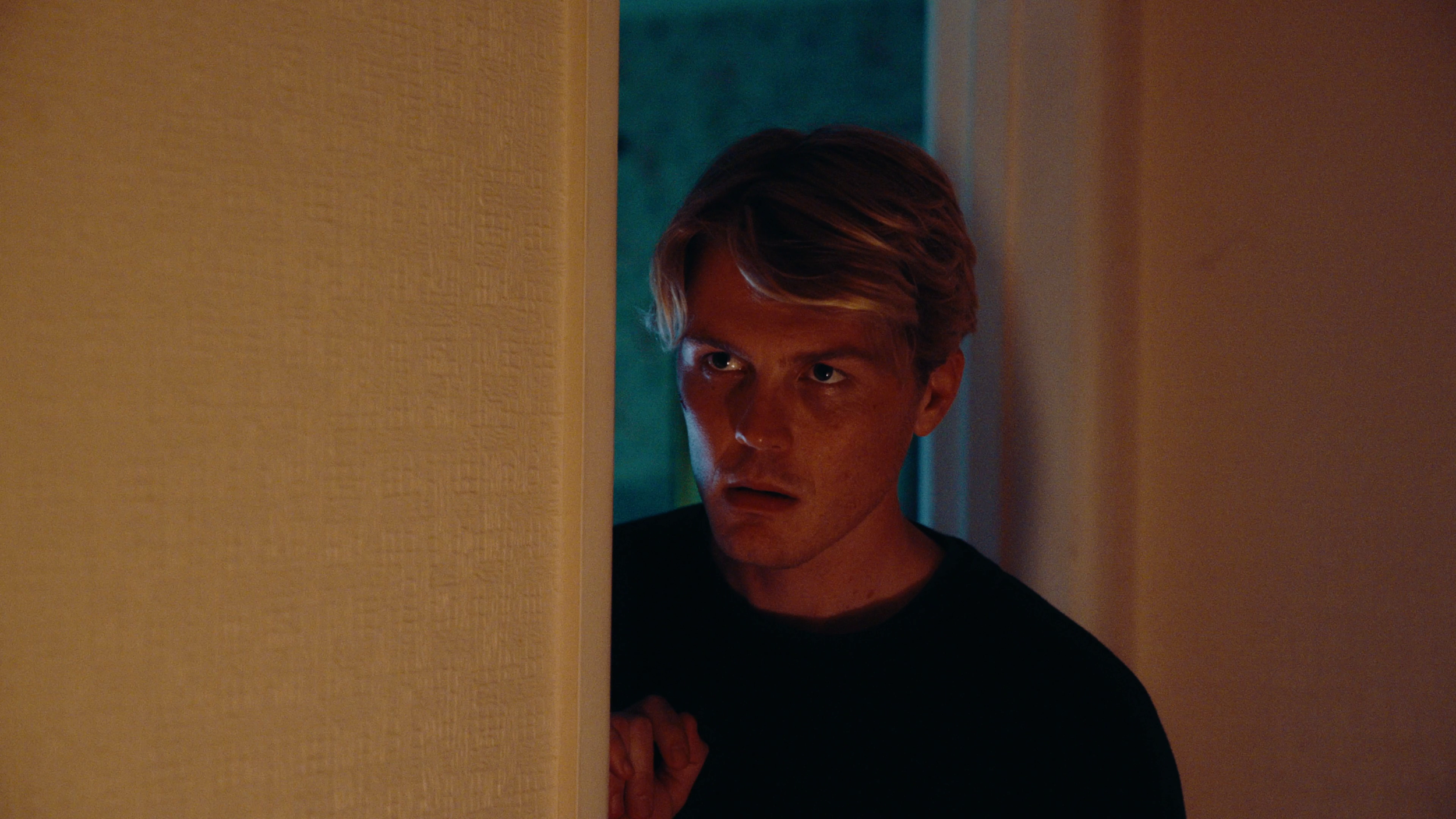 A young man with light brown hair peeks around a corner with a serious expression, holding the wall with one hand in a dimly lit room.