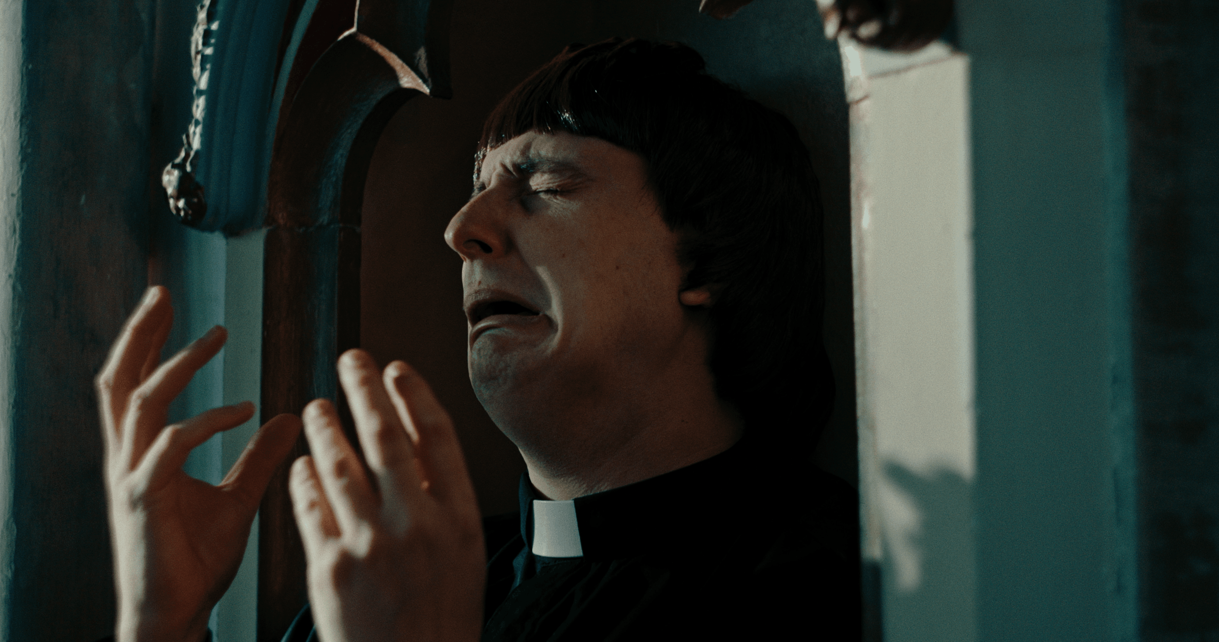 A person wearing a priest's collar with eyes closed, pressed against a wall and holding their hand up with fingers spread.