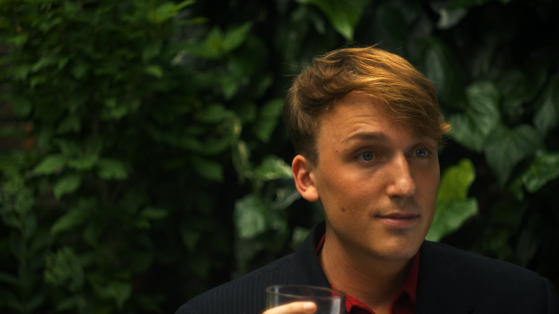 A young man with light brown wavy hair and blue eyes sitting in front of green foliage, holding a glass of a beverage.
