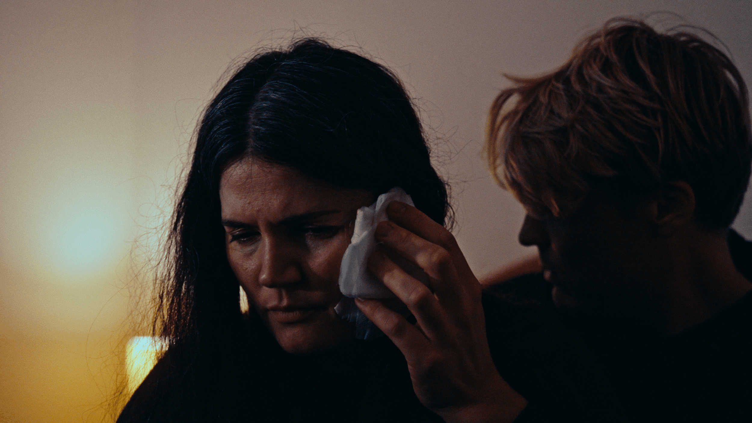 A woman with long dark hair is wiping tears from her face with a tissue while a man leans in close, offering comfort.