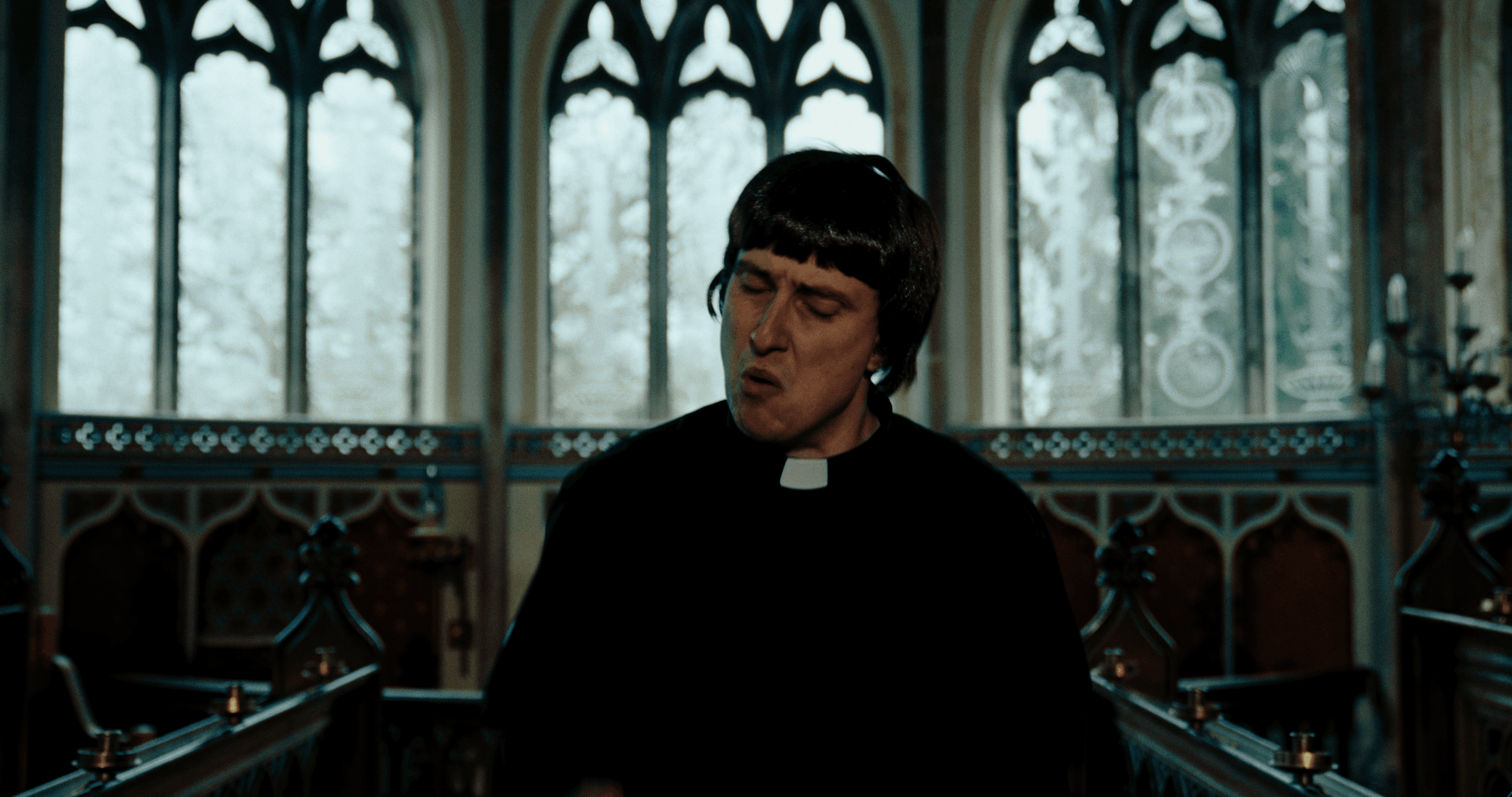 A man dressed as a priest in a church with stained glass windows in the background.