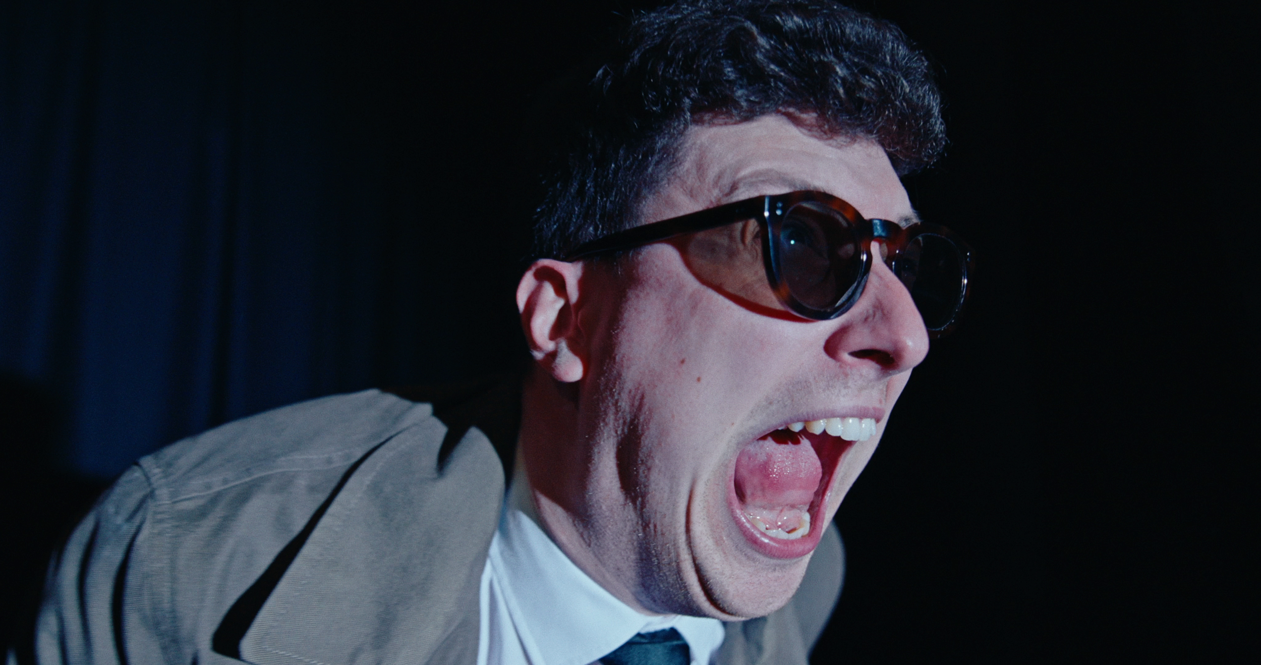 A man with curly hair and sunglasses is yelling with his mouth wide open, showing his teeth and tongue, in a dark setting.