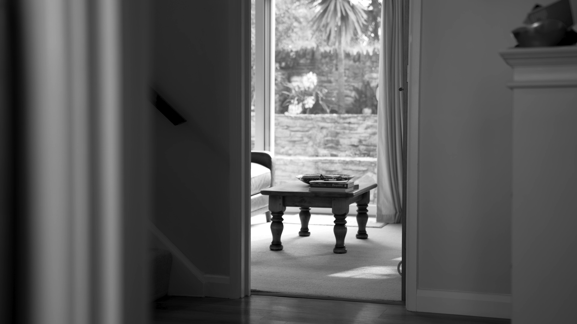 A black and white photo taken from a dark interior, looking through a doorway into a bright room with a window and curtain. There is a small wooden table with books and a tray on it, a chair beside it, and lush trees visible outside the window.
