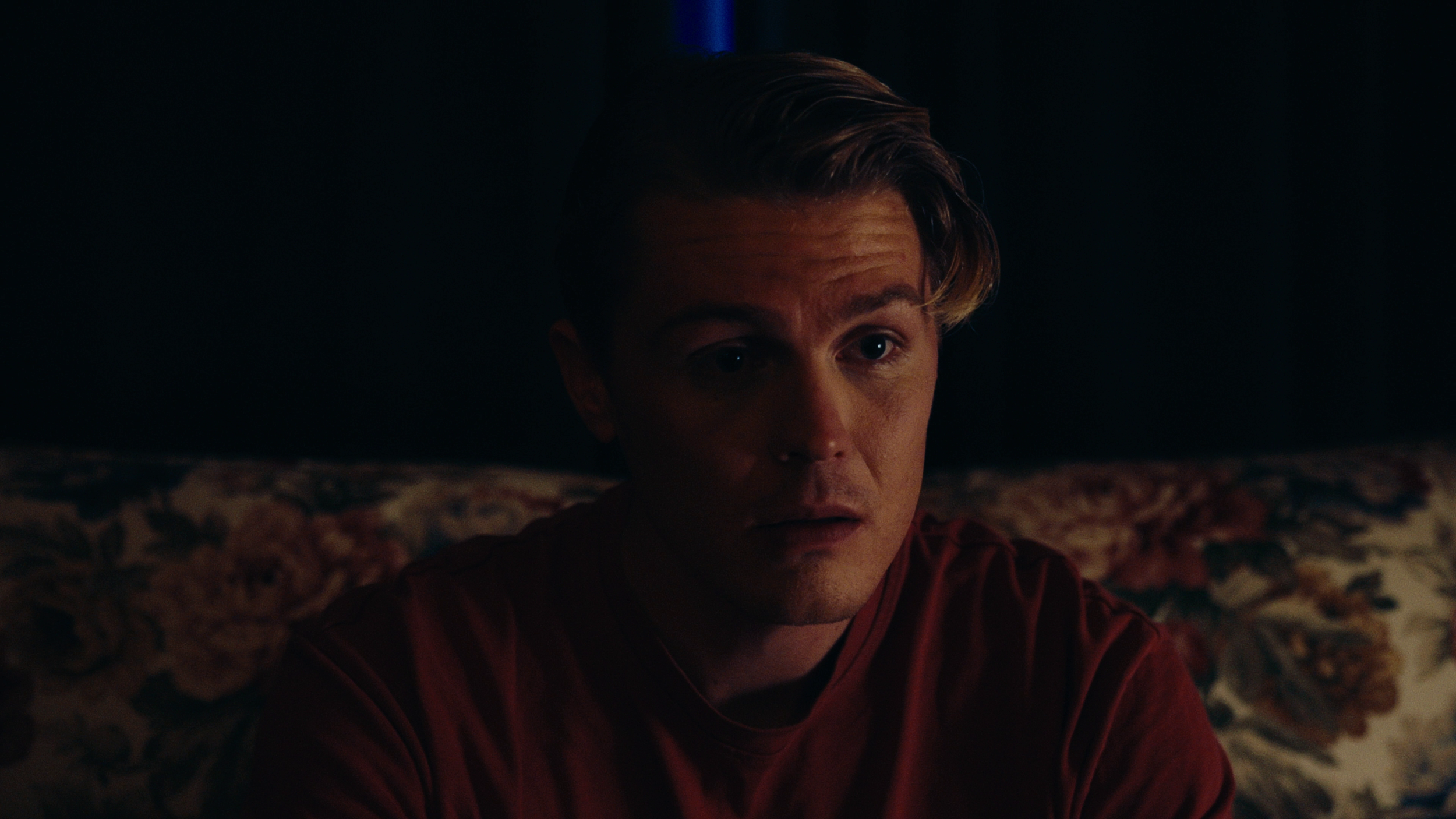 A young man with blond hair and a surprised or concerned expression, sitting on a floral-patterned couch in a dimly lit room.