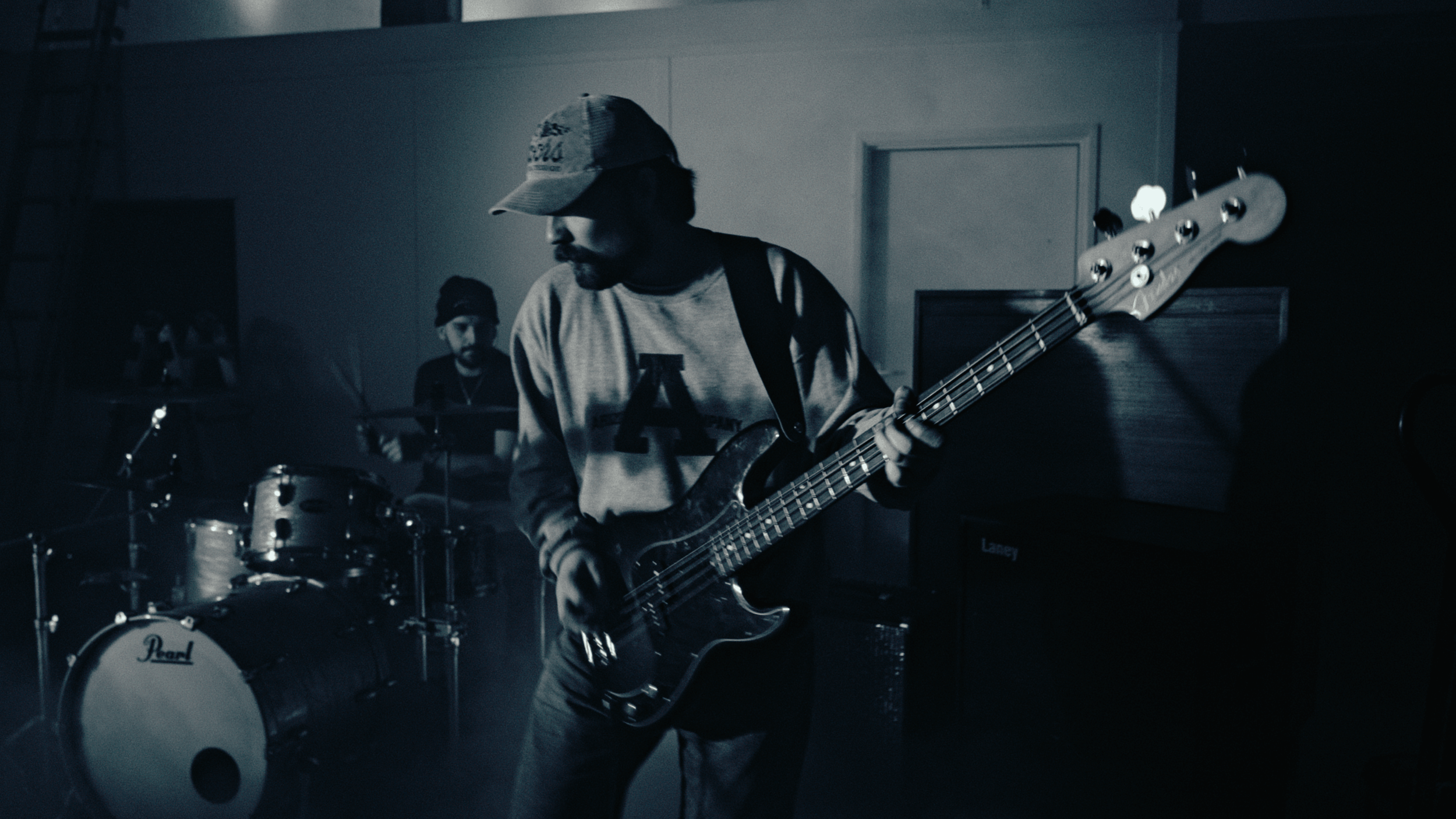 A musician wearing a baseball cap playing an electric bass guitar during a band rehearsal or performance, with drums visible in the background.
