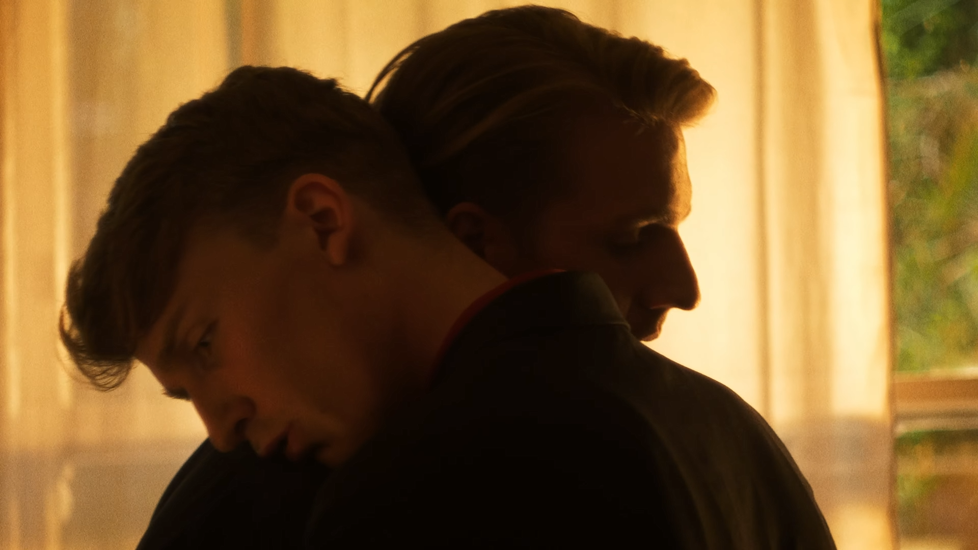Two young men hugging, with their faces close and eyes closed in a tender moment, in warm indoor lighting.