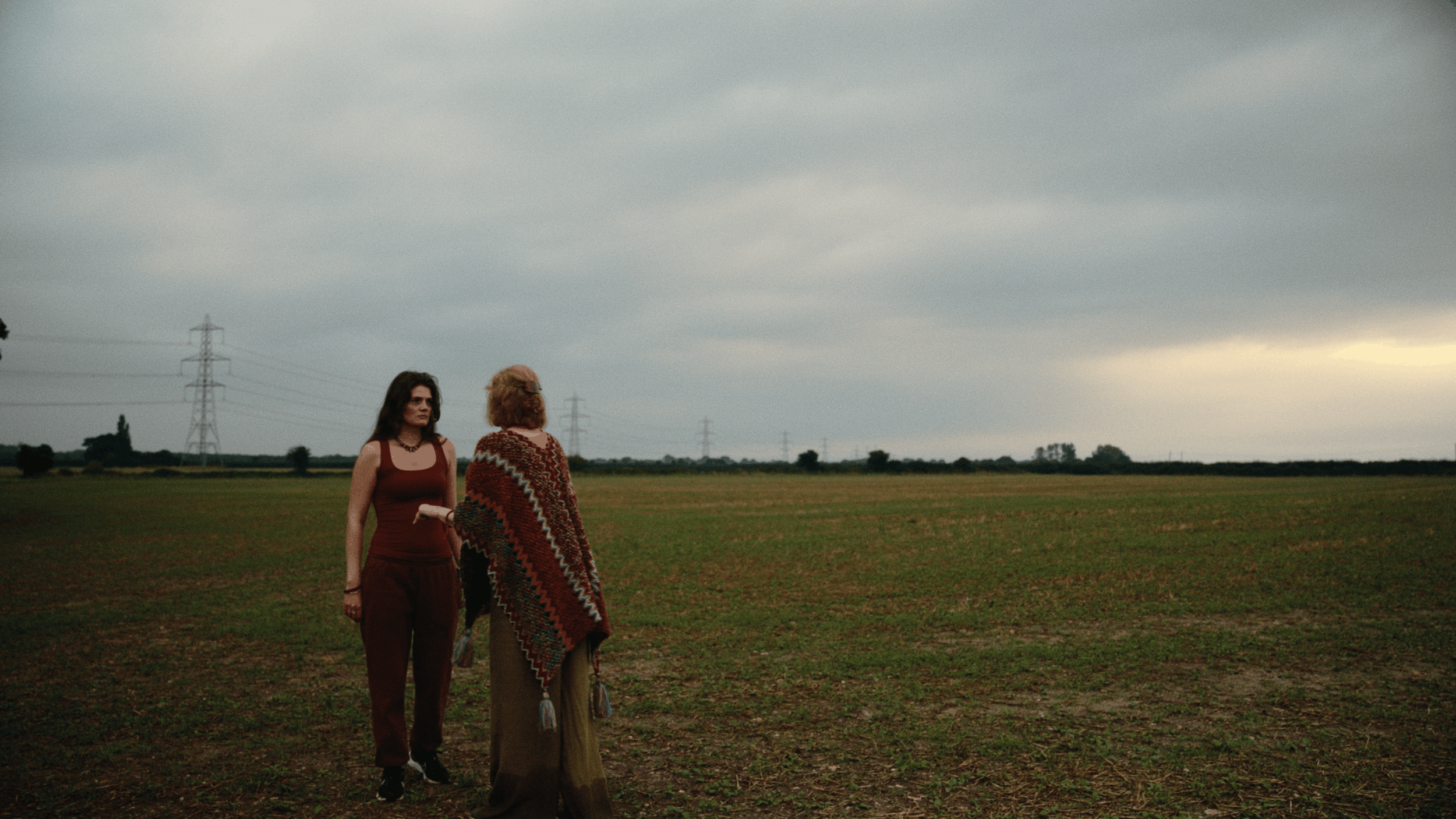 Two women standing in a vast field under a cloudy sky, with power lines in the background.