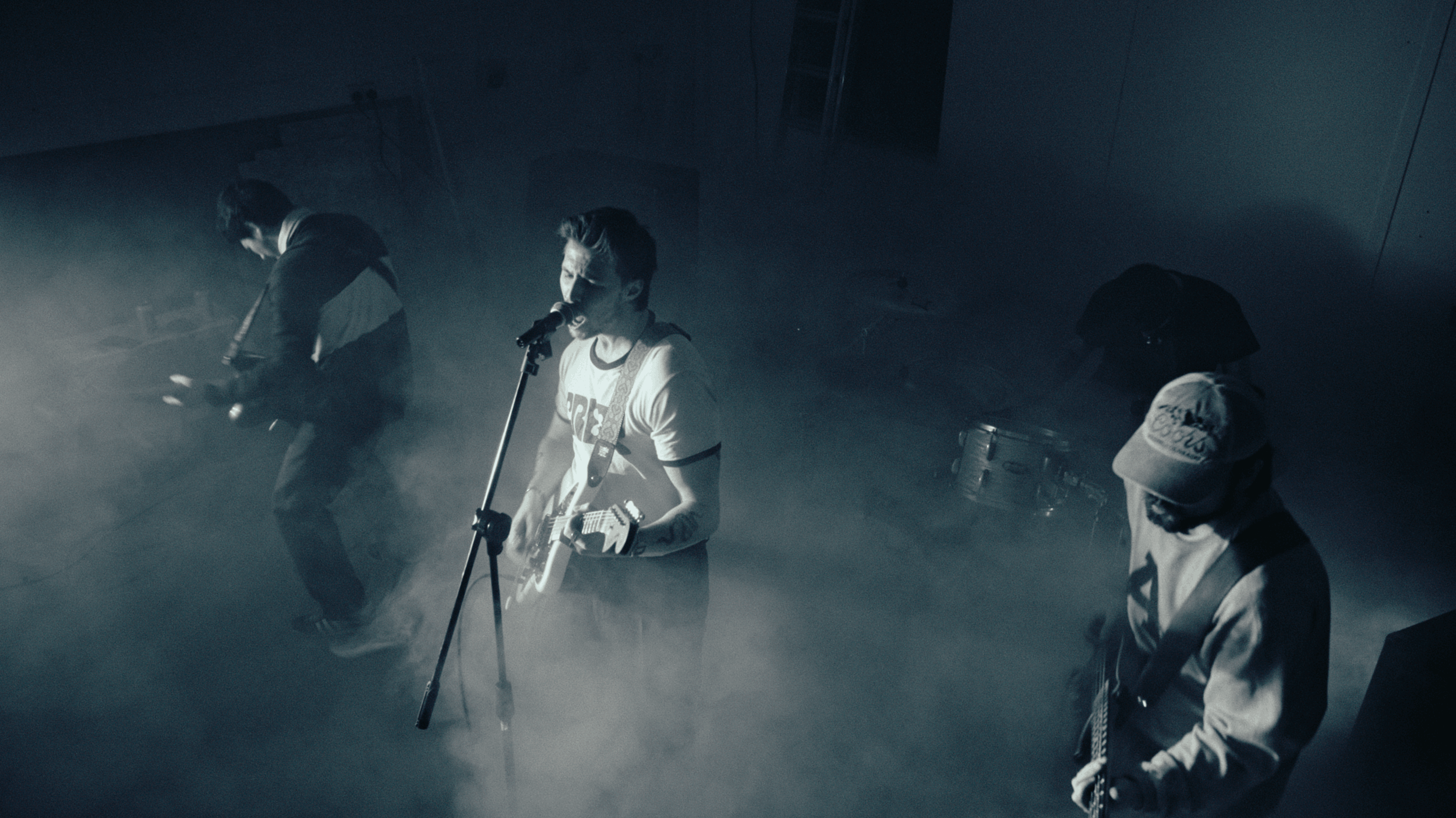 Four musicians performing on stage, one playing guitar and singing into a microphone, in a dark, smoky atmosphere.