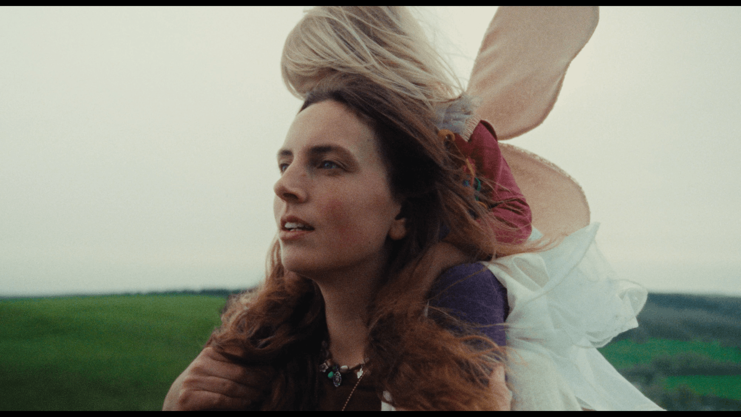 A woman with long red hair looking into the distance outdoors, with a man partially behind her carrying her on his back, set against a green field and overcast sky.