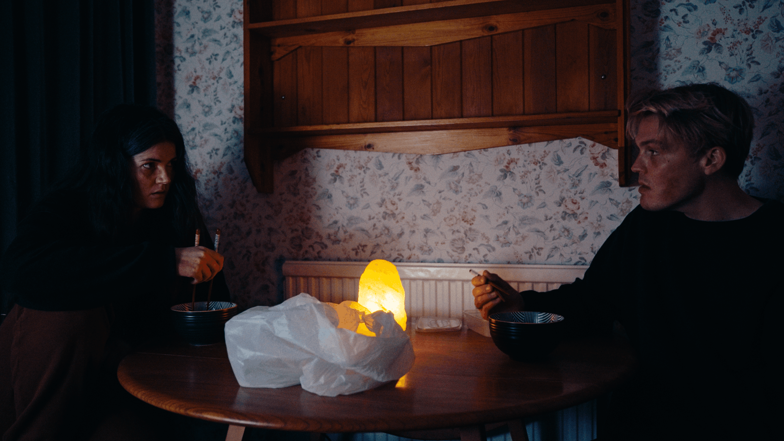 A dark room with two people sitting at a round wooden table, illuminated by a salt lamp. A woman with dark hair on the left and a man with blond hair on the right are facing each other, holding chopsticks, with bowls in front of them. The room has floral wallpaper and wooden shelves on the wall.