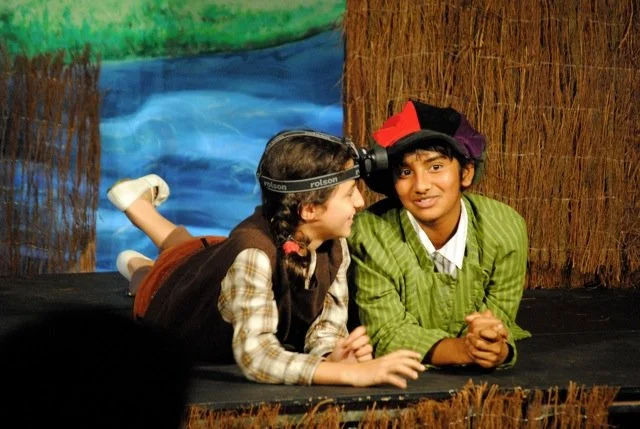 Two children in costume perform in a school play with a painted backdrop of water and trees, one wearing a headlamp and the other in a colorful hat.