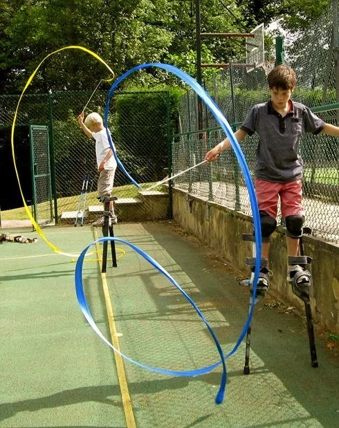 Two boys on stilts playing with hula hoops on an outdoor court surrounded by a chain-link fence, trees in the background.