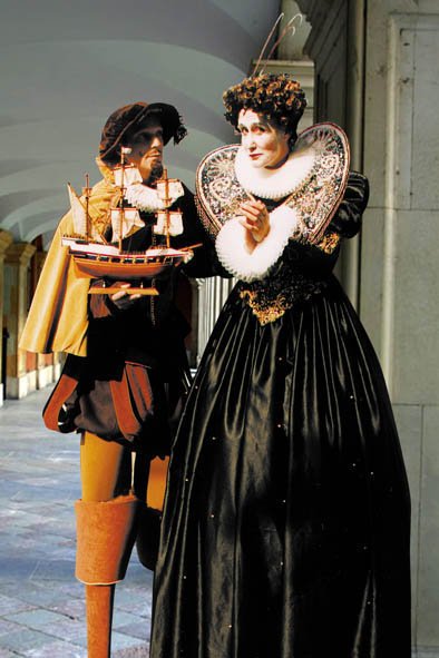 Two street performers dressed as historical figures in elaborate costumes, with one holding a ship model and the other standing with a hand near her face.