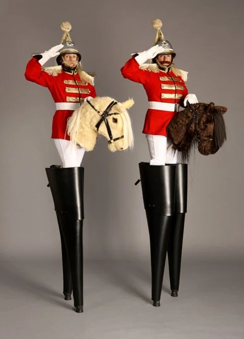 Two women dressed as toy soldiers on stilts, each riding a toy horse, saluting with their right hands, against a plain gray background.