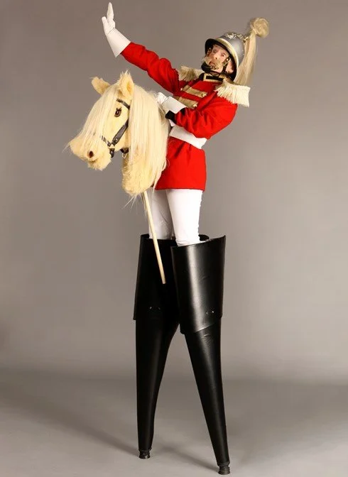 A person dressed as a nutcracker soldier, riding a large puppet horse with a white mane, wearing a red uniform with white trousers, a silver helmet, and tall black boots, standing on a pair of large black stilts.