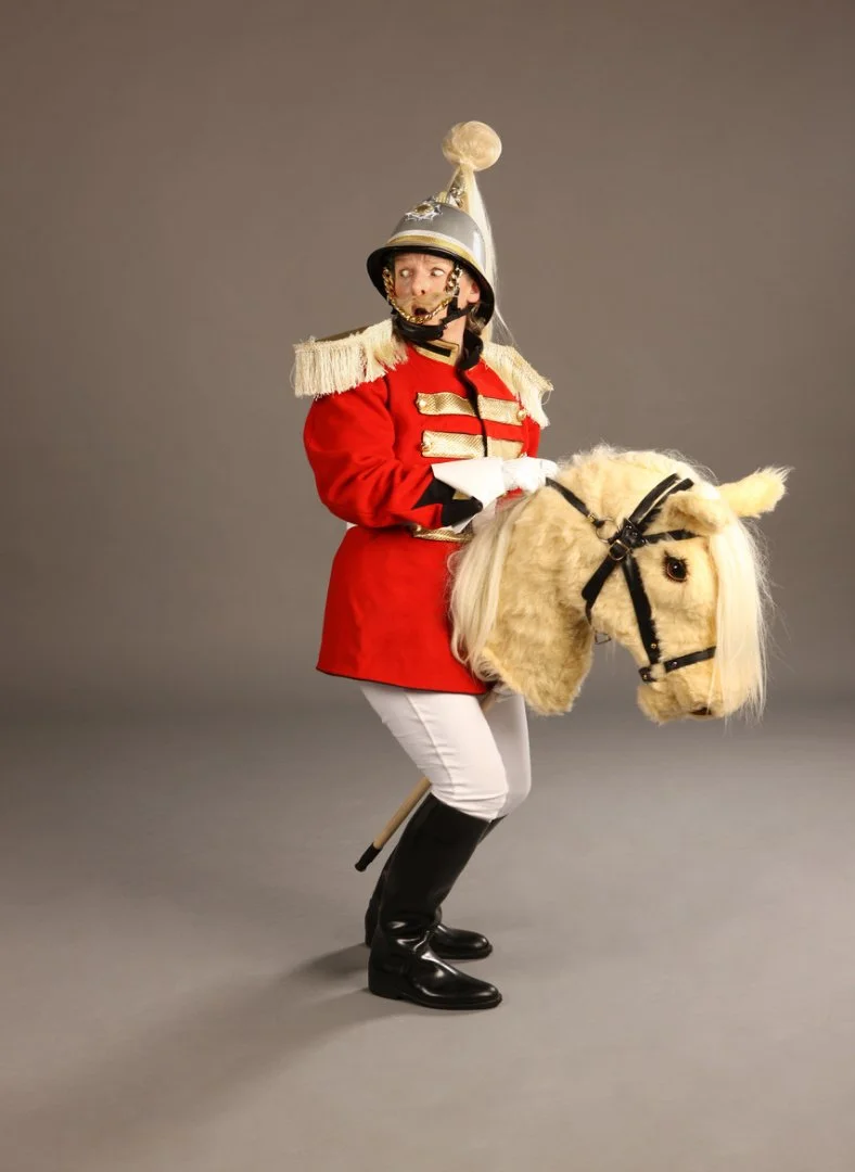 Person dressed as a toy soldier riding a plush hobby horse against a plain gray backdrop.
