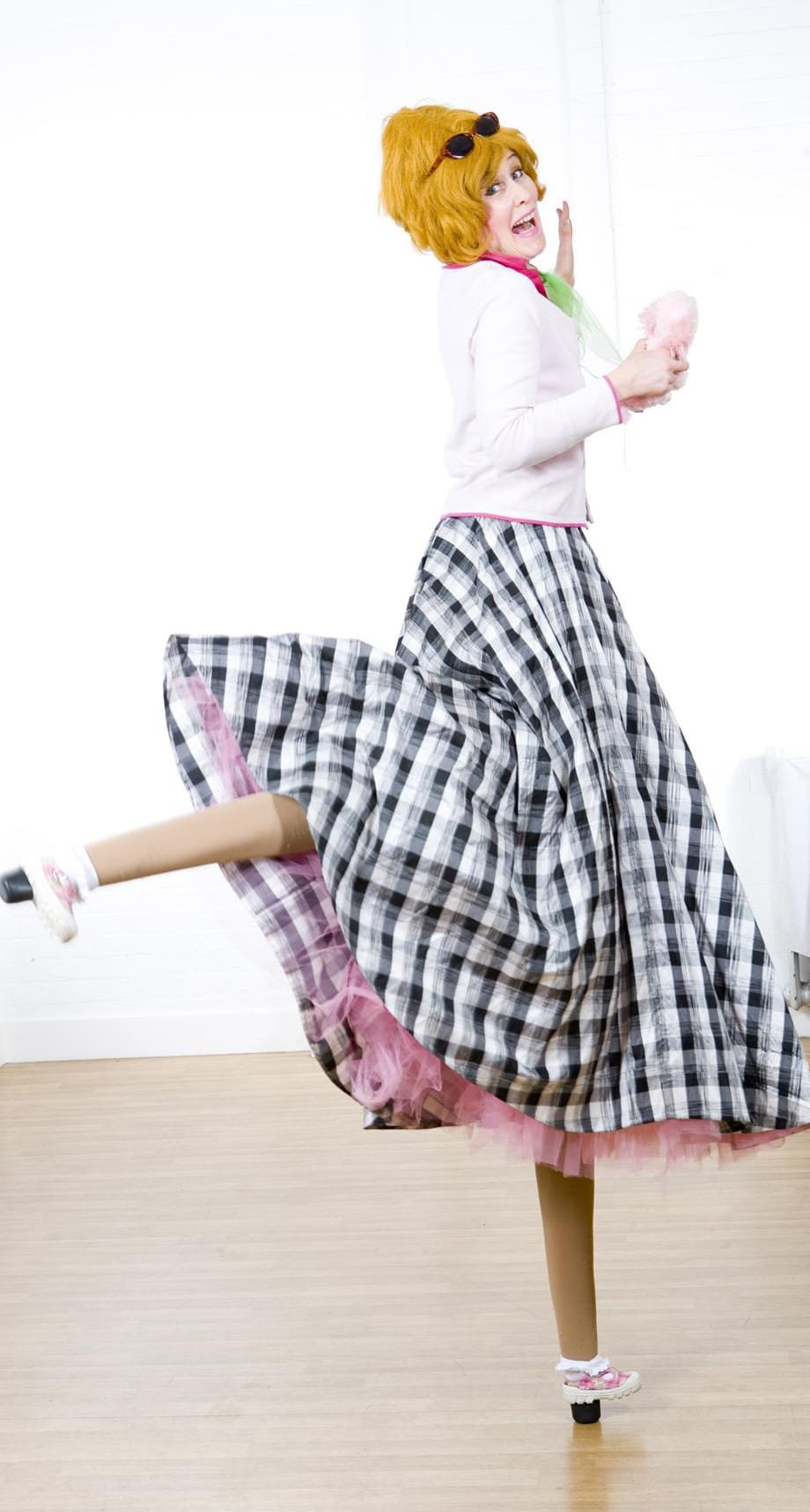 Person in colorful costume with red wig, plaid skirt, white top, orange tights, and roller skate, striking a playful pose against a white background.