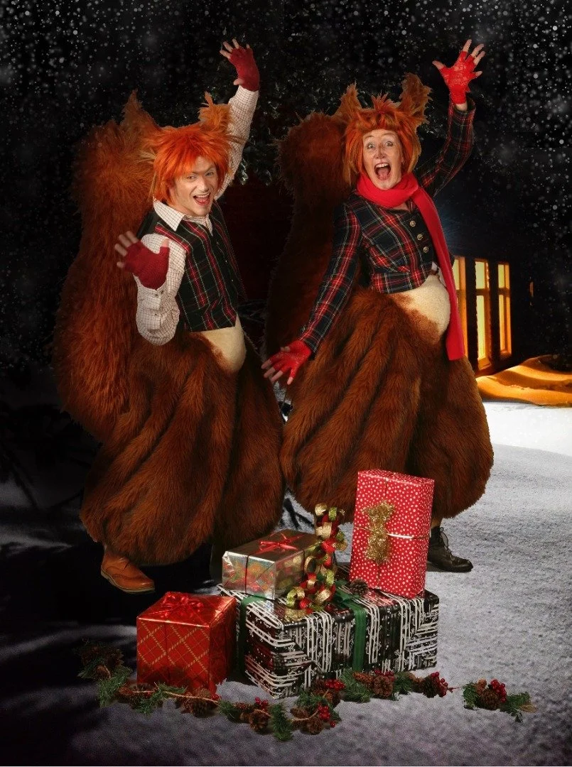 Two people dressed as foxes in festive Christmas costumes with fur tails, red scarves, and plaid jackets, standing on a snowy surface surrounded by wrapped presents and holiday decorations.