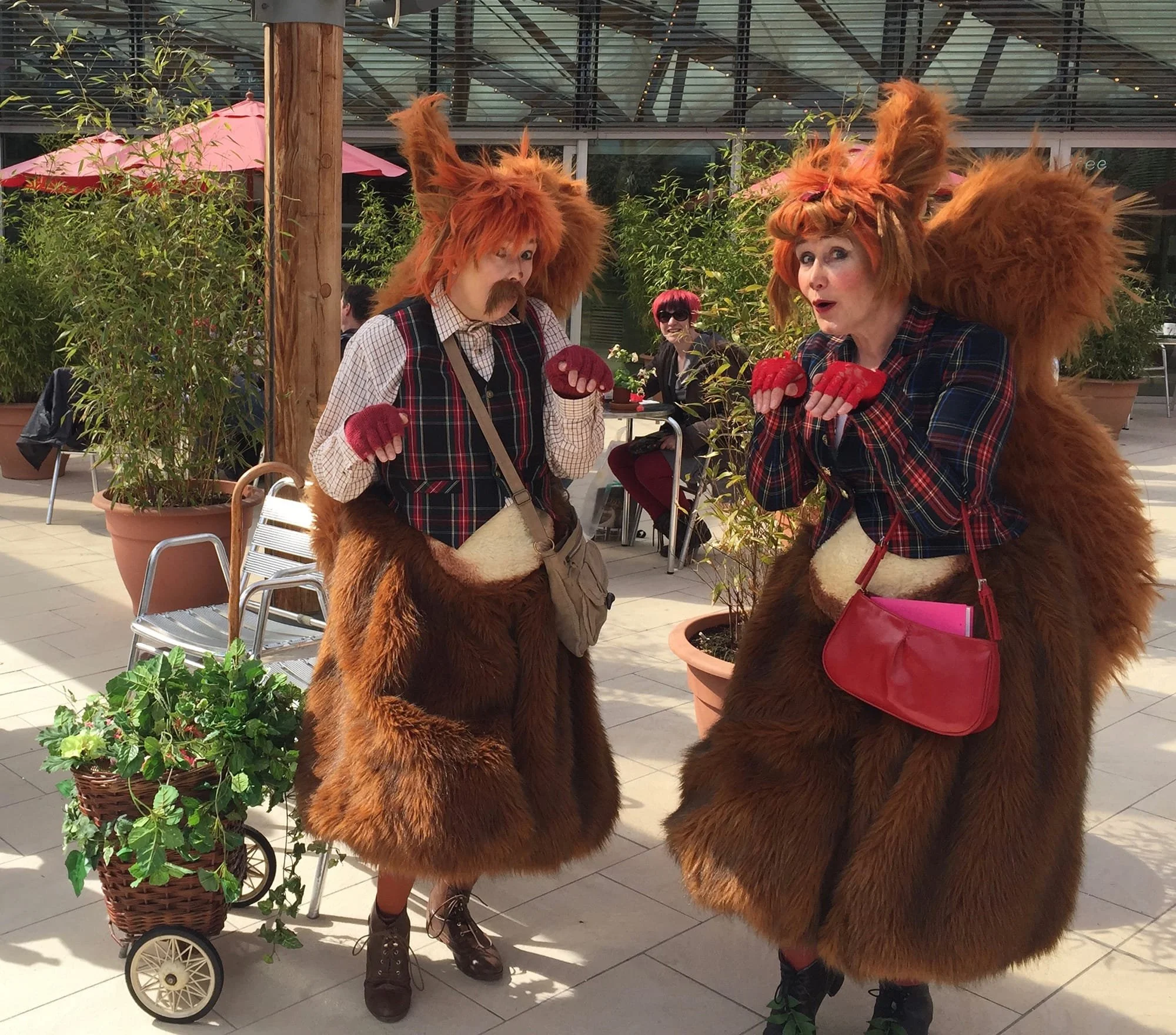 Two women dressed as lions with large, furry, orange manes and lion costumes are engaging in a playful interaction in an outdoor patio setting. One woman has a mustache and is holding her hands up, while the other is gesturing with her hands. In the 