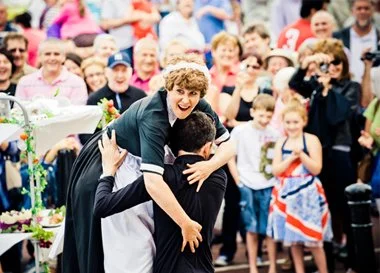Two men are dancing and embracing at an outdoor event with a crowd of smiling people watching.