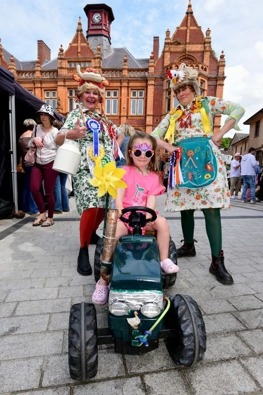 Two clowns dressed in colorful costumes and hats with animal theme accessories standing next to a young girl sitting on a green toy tractor at a festival or fair, with a historic brick building in the background.