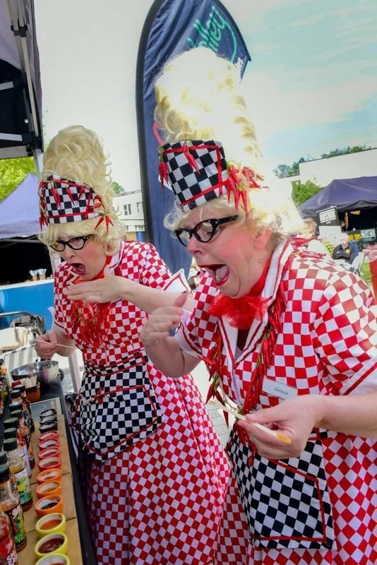 Two women dressed as chefs in red and white checkered uniforms, with large curly blonde wigs, glasses, and checkered hats, tasting or sampling food at an outdoor event with tents and banners in the background.