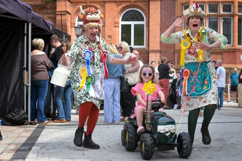 Two women dressed as clowns with colorful ribbons and costumes, entertaining a young girl riding a small tractor toy at an outdoor event. The woman on the left wears a dragon hat, and the woman on the right has a hat with a chicken and red, yellow, a
