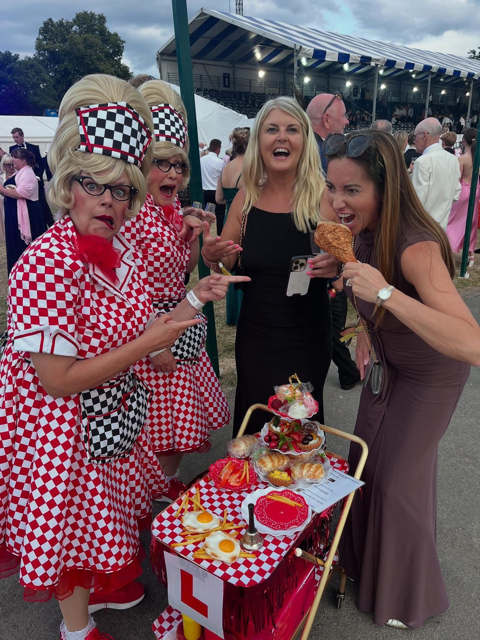 Four women at an outdoor event, with two dressed in matching red and white checkered dresses and headbands, and two in modern dresses. They are gathered around a cart with colorful cupcakes and desserts, with one woman eating a corn dog. The backgrou