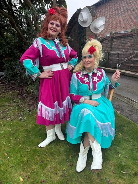 Two women dressed in colorful vintage 1950s-style dresses with star and fringe details, one standing and one sitting outside, with garden and brick wall background.