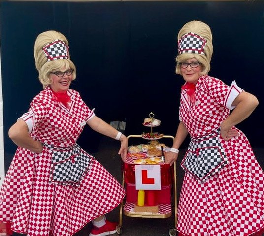 Two women in vintage waitress costumes with red and white checkered dresses and matching hats stand on either side of a red cart with an 'L' license plate, black background.