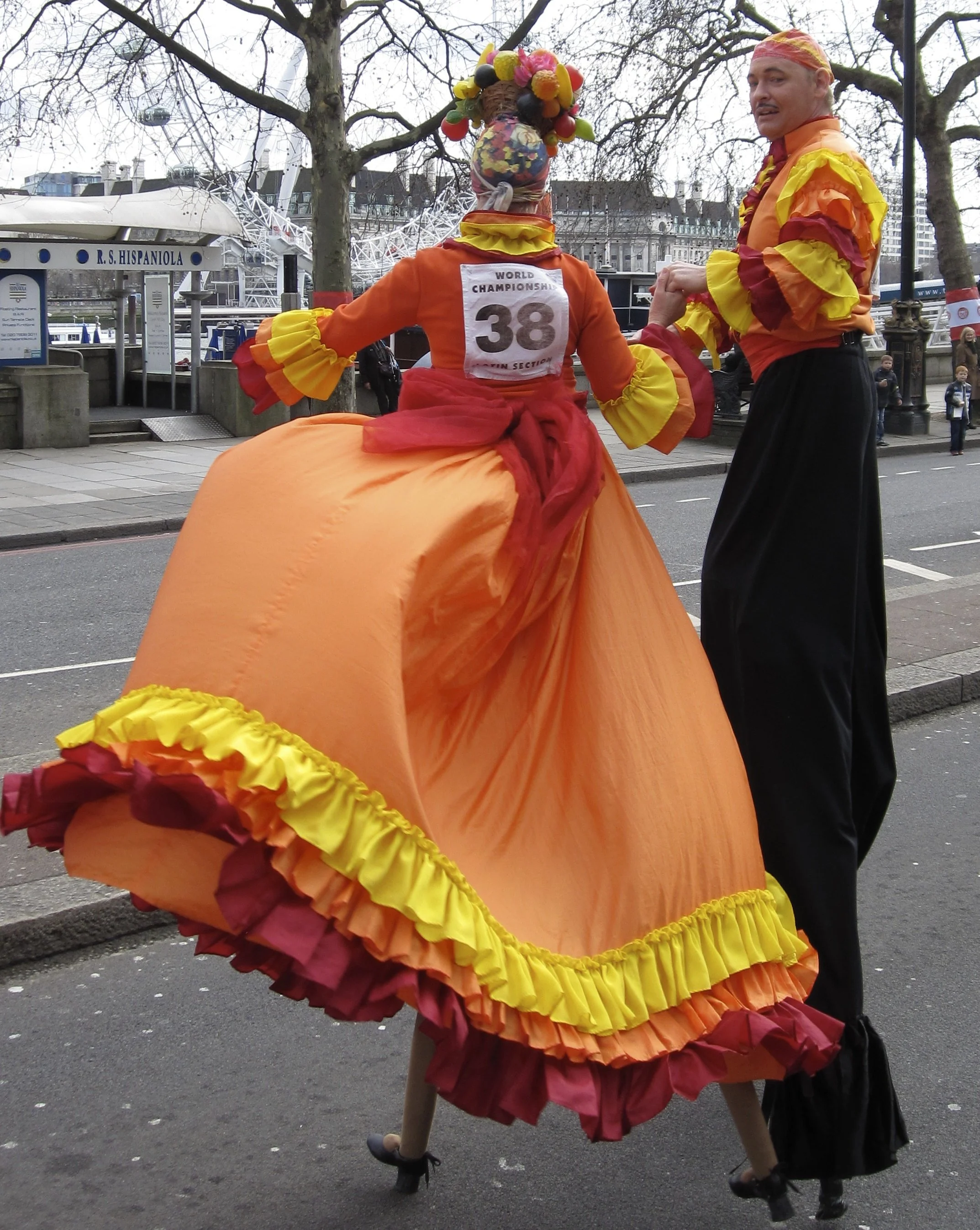 Two dancers in colorful, floral costumes performing outdoors on a city street with leafless trees and buildings in the background.