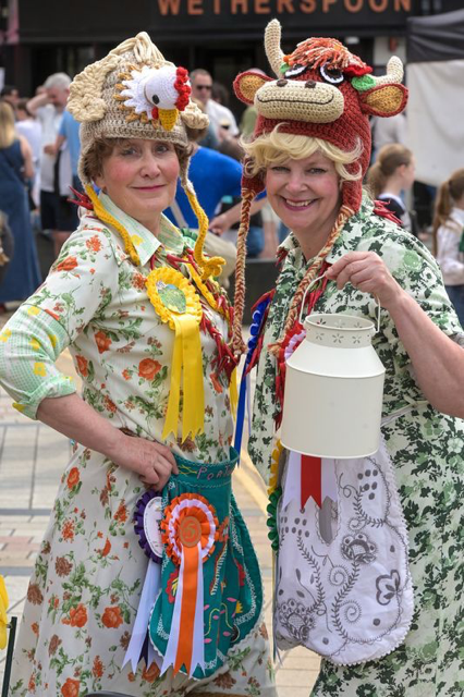Two women in festive costumes at a fair, wearing knitted animal hats resembling a rooster and a cow, with ribbons and minutes accessories, in an outdoor setting with people in the background.