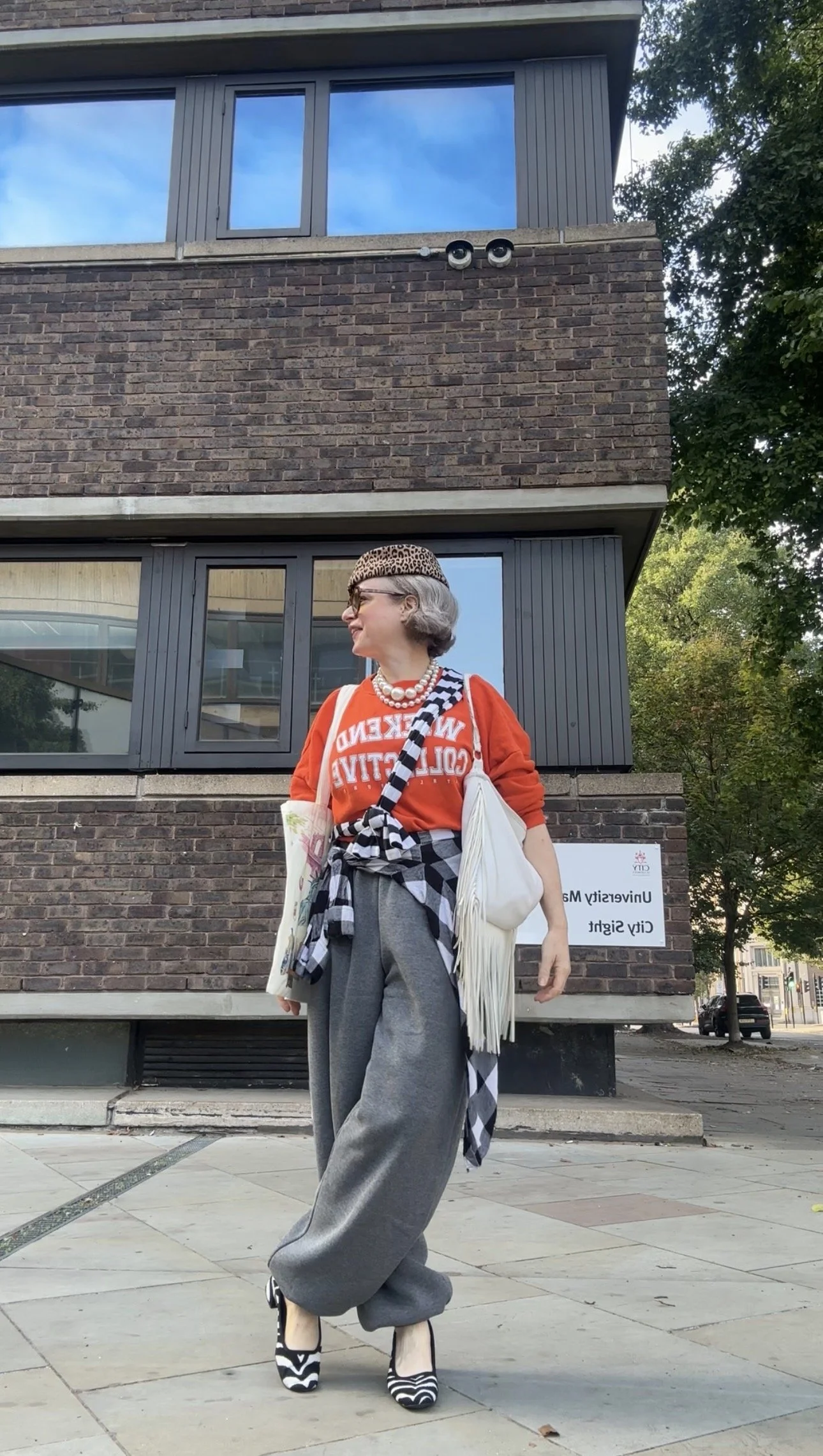 Woman over 40 outside in a streetwear vibe layered outfit grey tracksuit bottoms, orange sweatshirt, leopard pill box hat, zebra shoes and black and white clothes as accessories 