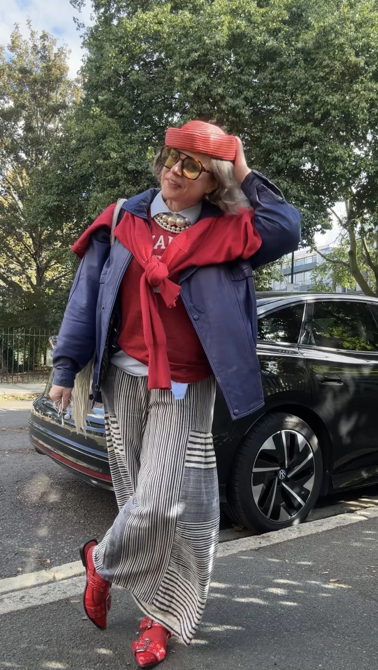 woman in layered outfit, blue leather jacket, red pillbox hat, stripy wide leg trousers, button down and sports vest with pearls, showing cardigan worn as a scarf layer to add outfit cohesio