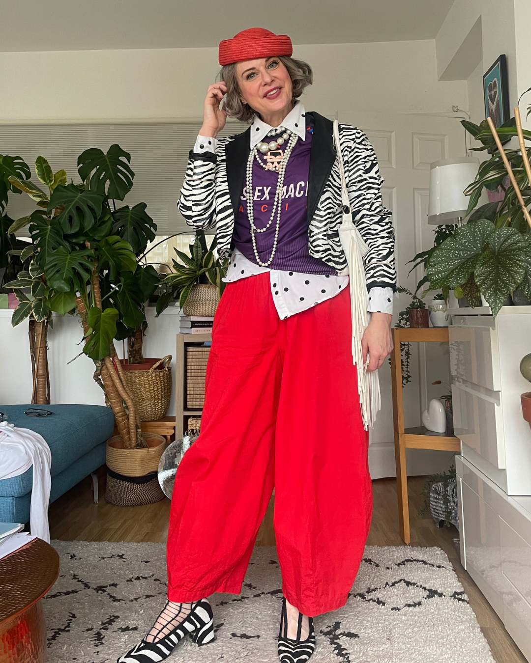 Full layered autumn outfit: woman over 40 in red pillbox beret  zebra print jacket over purple sweatshirt and white polka dot shirt, pearls, red balloon-leg trousers.