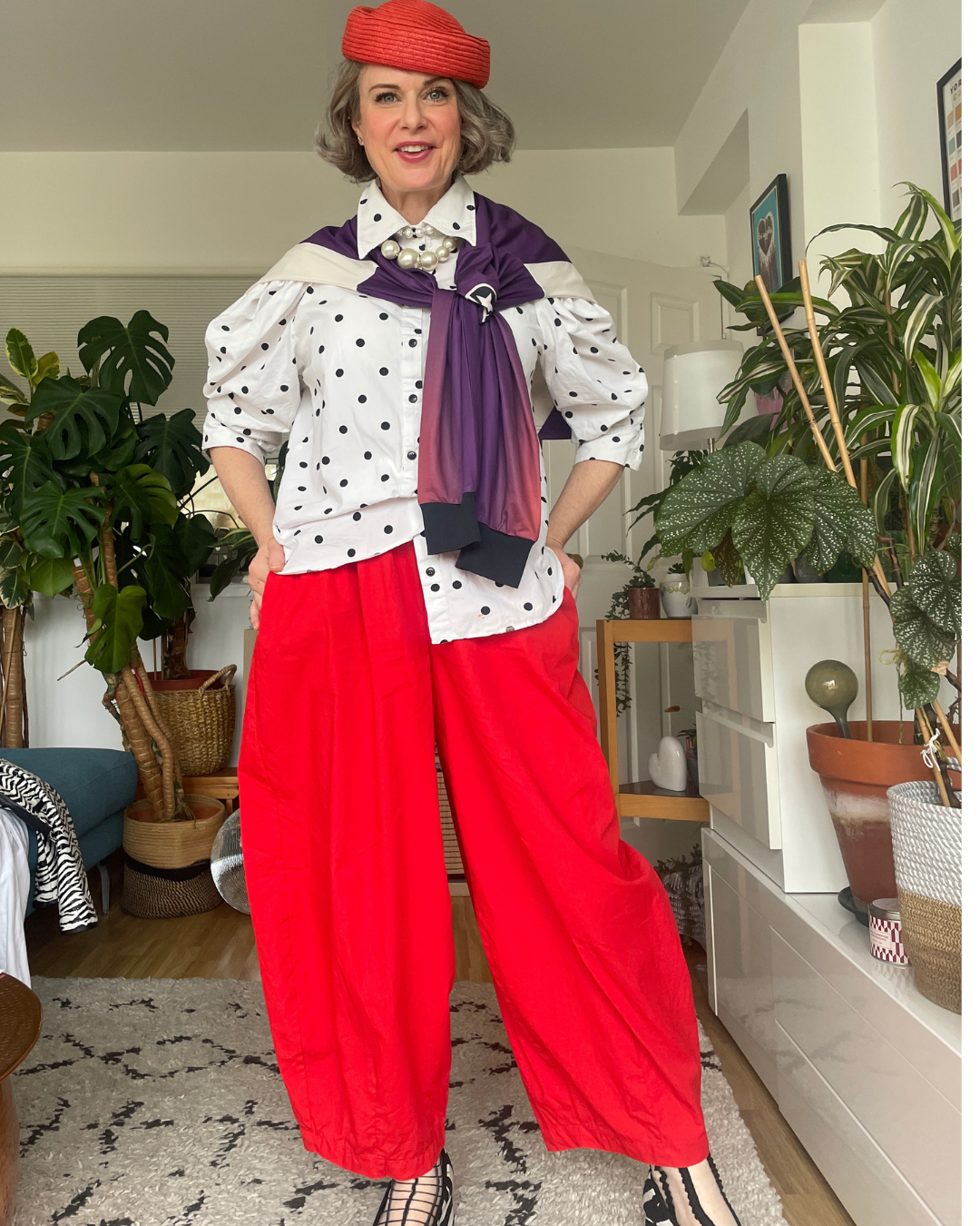 Purple long sleeve top taken off and worn as an accessory, slung over the shoulders like a scarf on a white polka dot puff-sleeve shirt with red trousers.