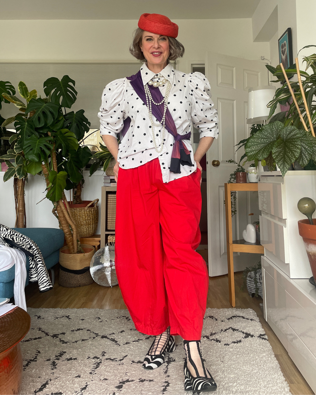 Purple long sleeve top restyled, tied across the body over a white polka dot shirt, with pearls, red pillbox beret and red wide-leg trousers.