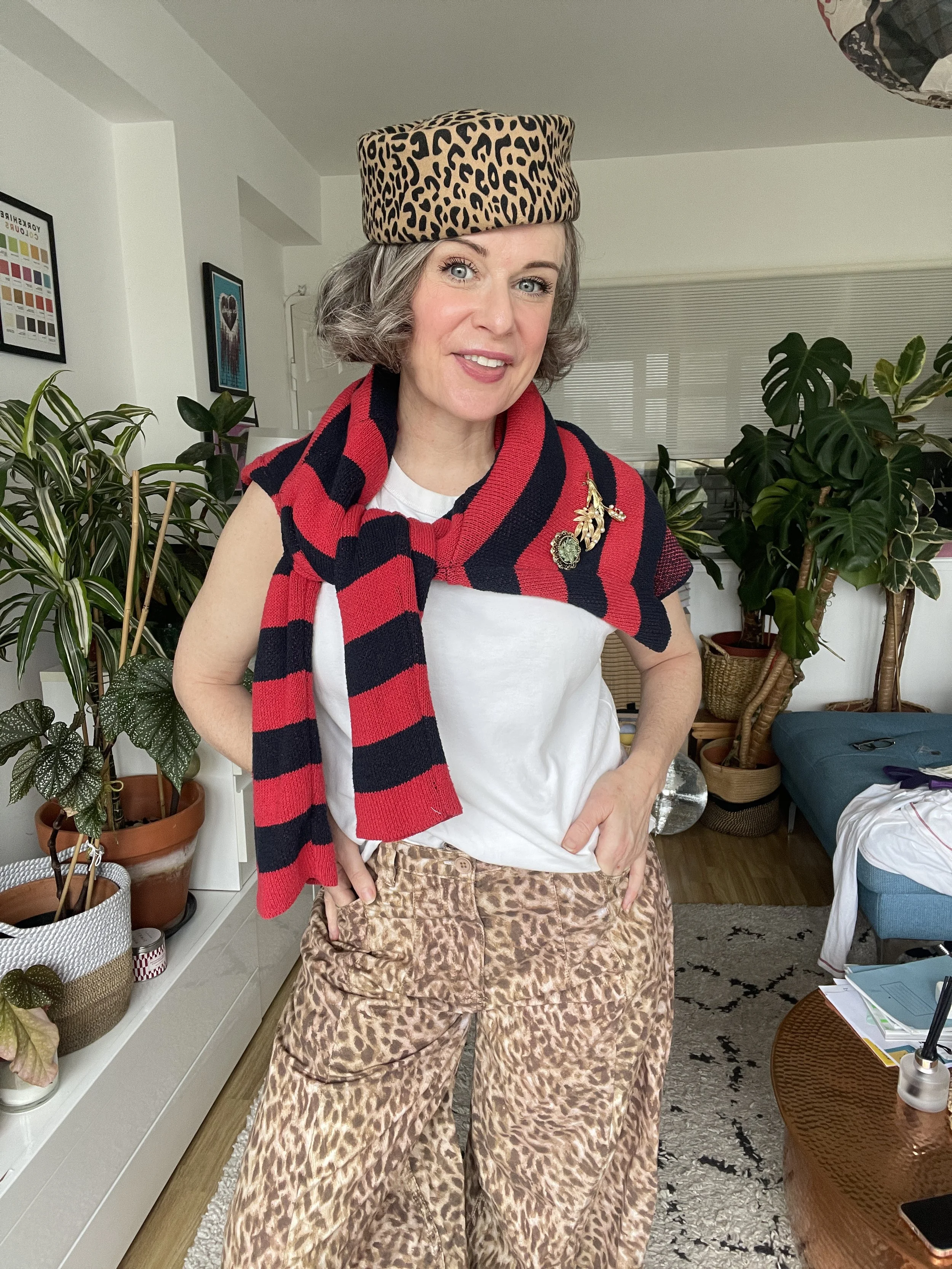 Woman over 40 showing a cardigan used as cool shawl  layer with brooches, she is wearing leopard hat and trousers  and the cardigan is black and red stripes