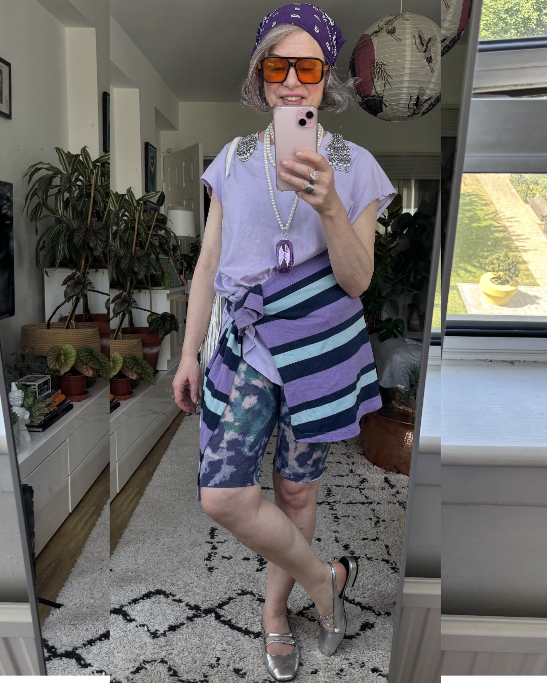 Creative layered outfit over 40 — lilac tee, striped skirt tied at waist, tie-dye bike shorts, purple bandana, silver Mary Janes — women's style over 40