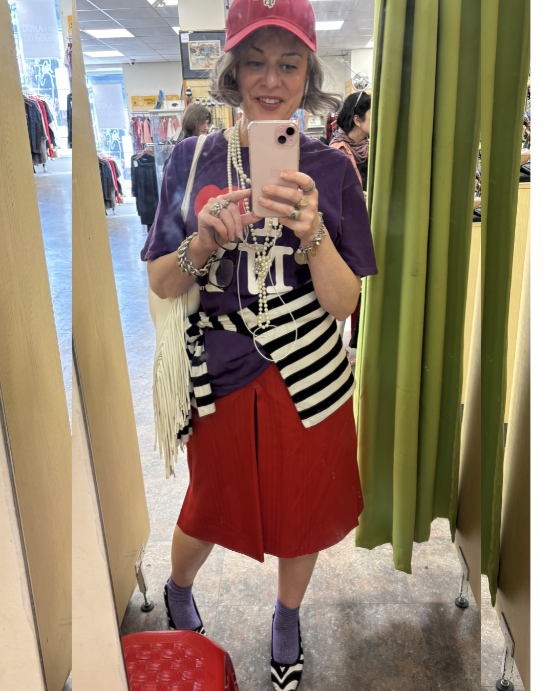Outfit  photo in a Thrift store of woman stylist who is over 40 — purple graphic tee, striped shirt, red midi skirt, red cap, pearl necklace, fringe bag — creative women's style over 40
