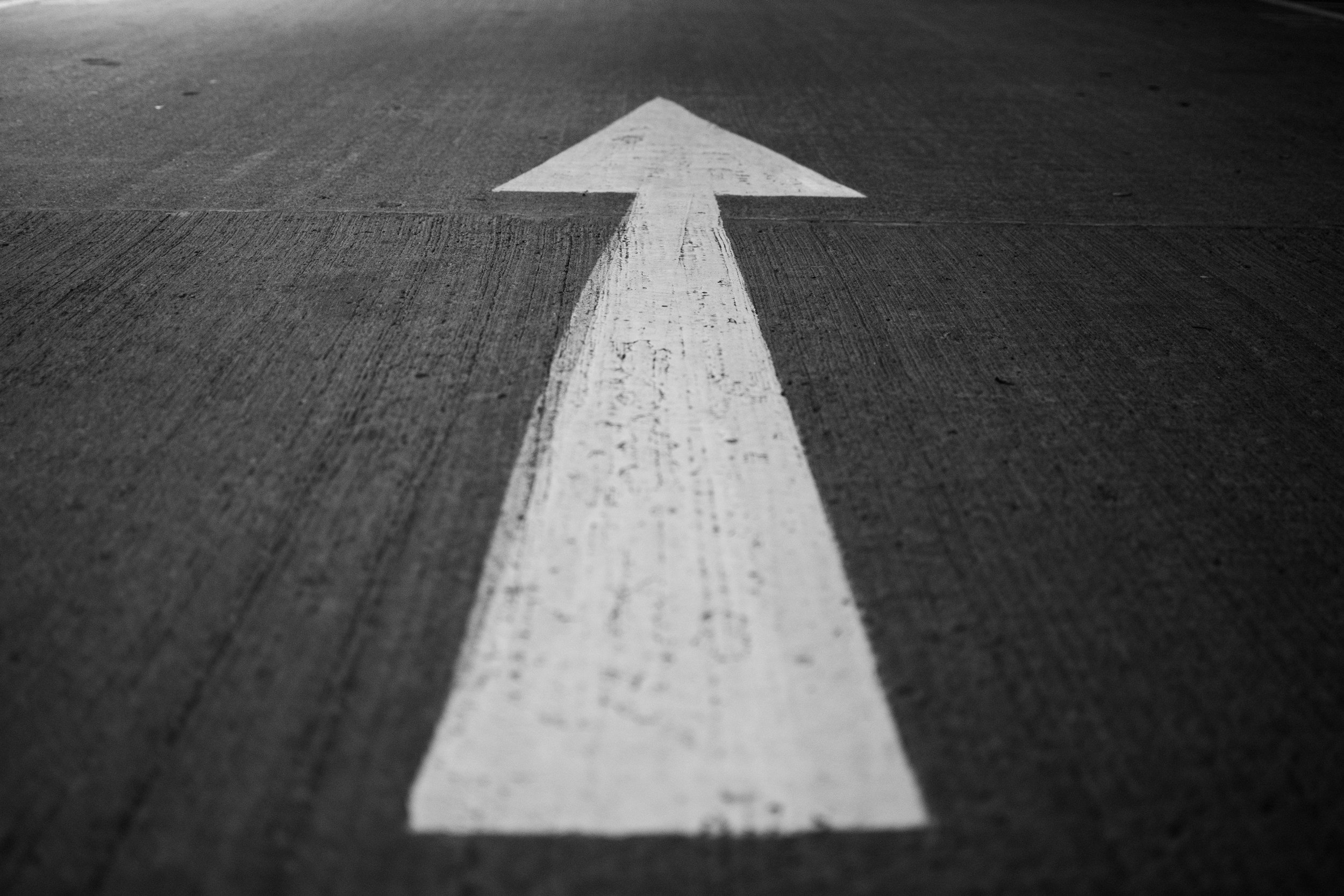 Black and white photo of an arrow painted on a concrete surface pointing forward.