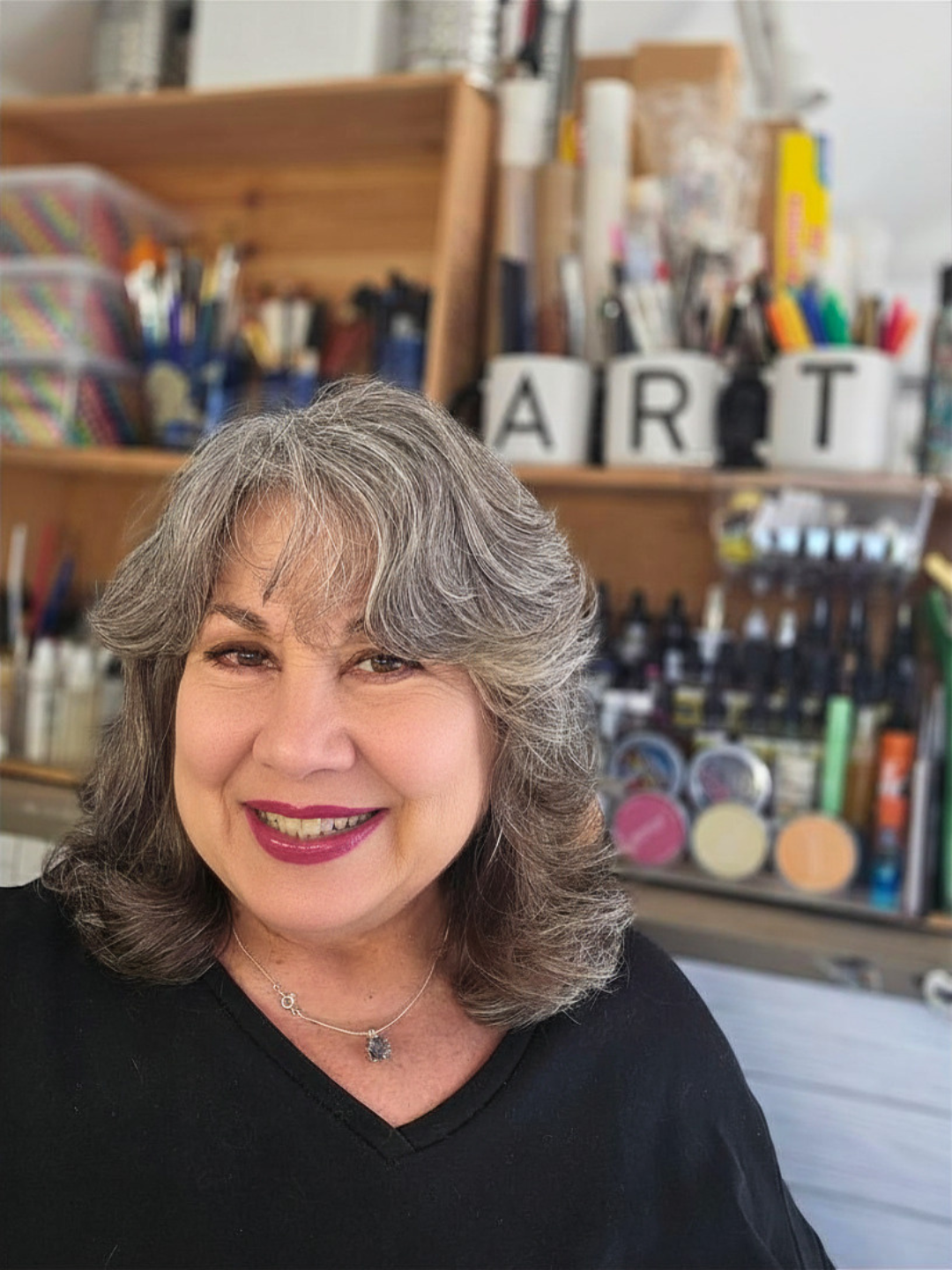 Lorraine Rimmelin with gray curly hair wearing a black top and a necklace, in a watercolor art studio with shelves of art supplies and letters spelling art in the background.